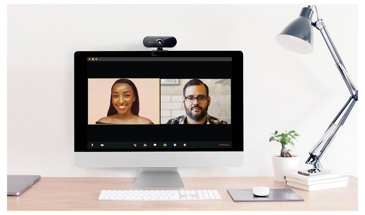 Ekran komputera wyświetlający rozmowę wideo z dwoma uczestnikami; jedna osoba po lewej stronie, a druga po prawej, na biurku z książkami i lampą.