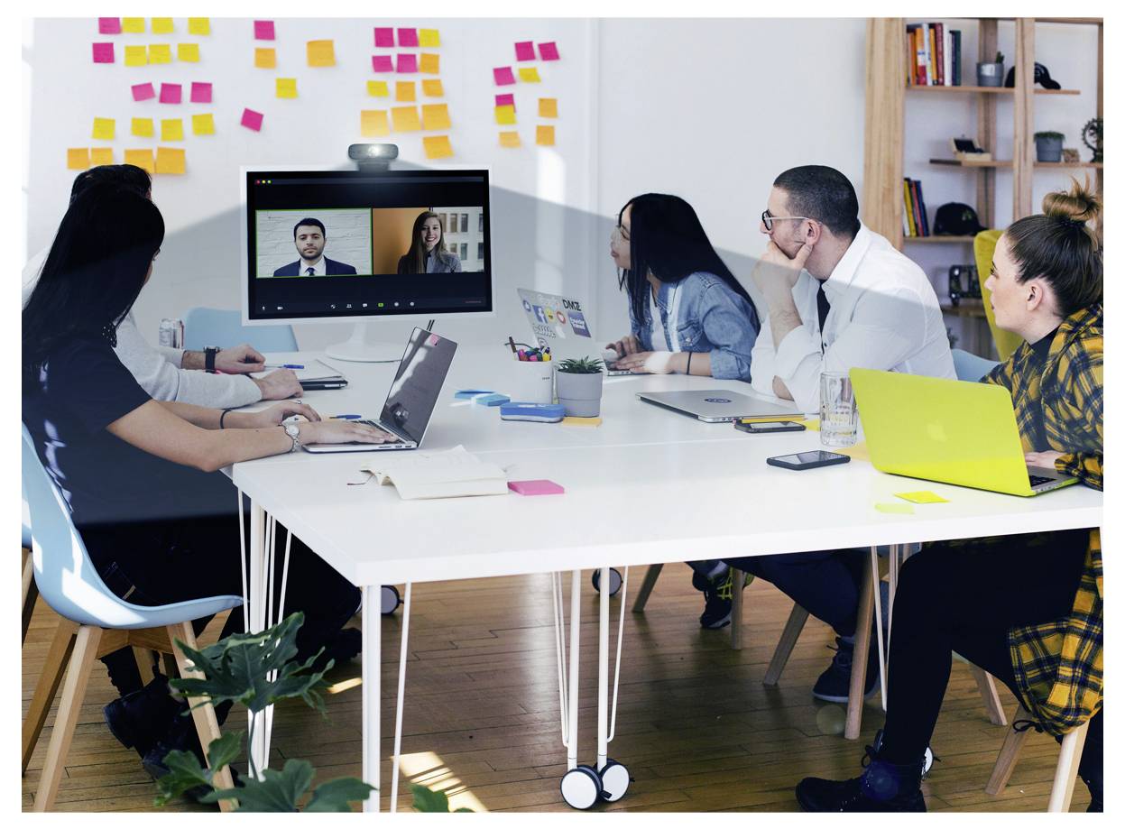 Zróżnicowana grupa ludzi siedzi wokół stołu w sali konferencyjnej, uczestnicząc w zdalnym spotkaniu wideo wyświetlanym na ekranie.