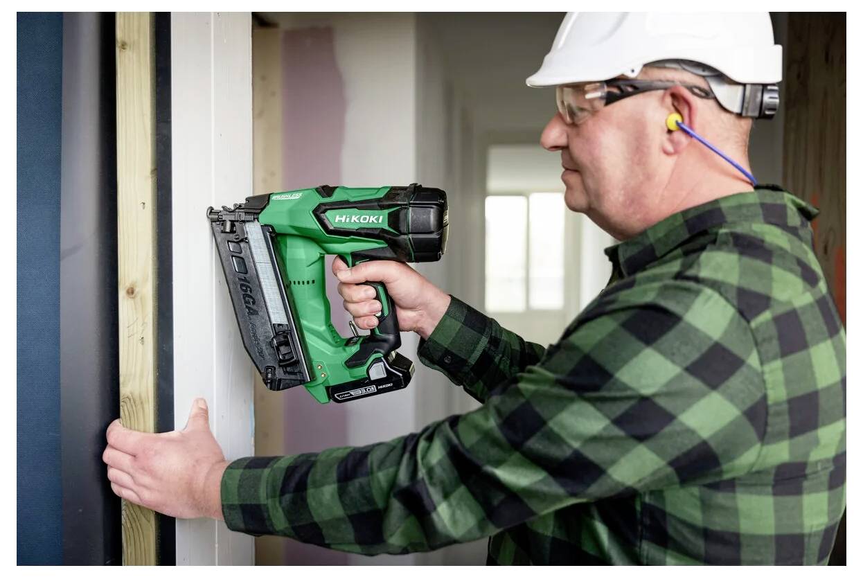 Osoba w kasku i ochronnikach słuchu używa zielonego gwoździarka do przymocowania drewnianej belki w pomieszczeniu.