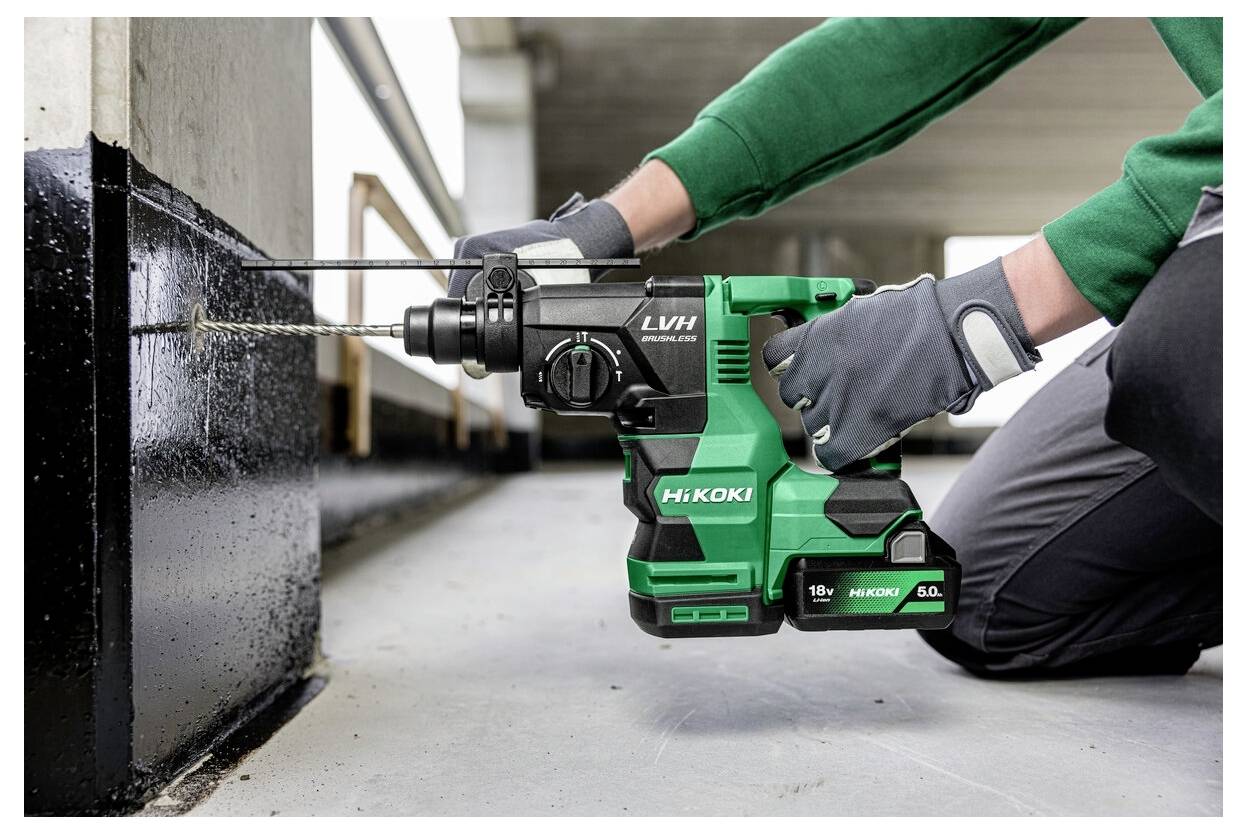 Osoba w szarych rękawicach używa zielonej i czarnej wiertarki do wykonania otworu w czarnej metalowej powierzchni, co wskazuje na działalność budowlaną.