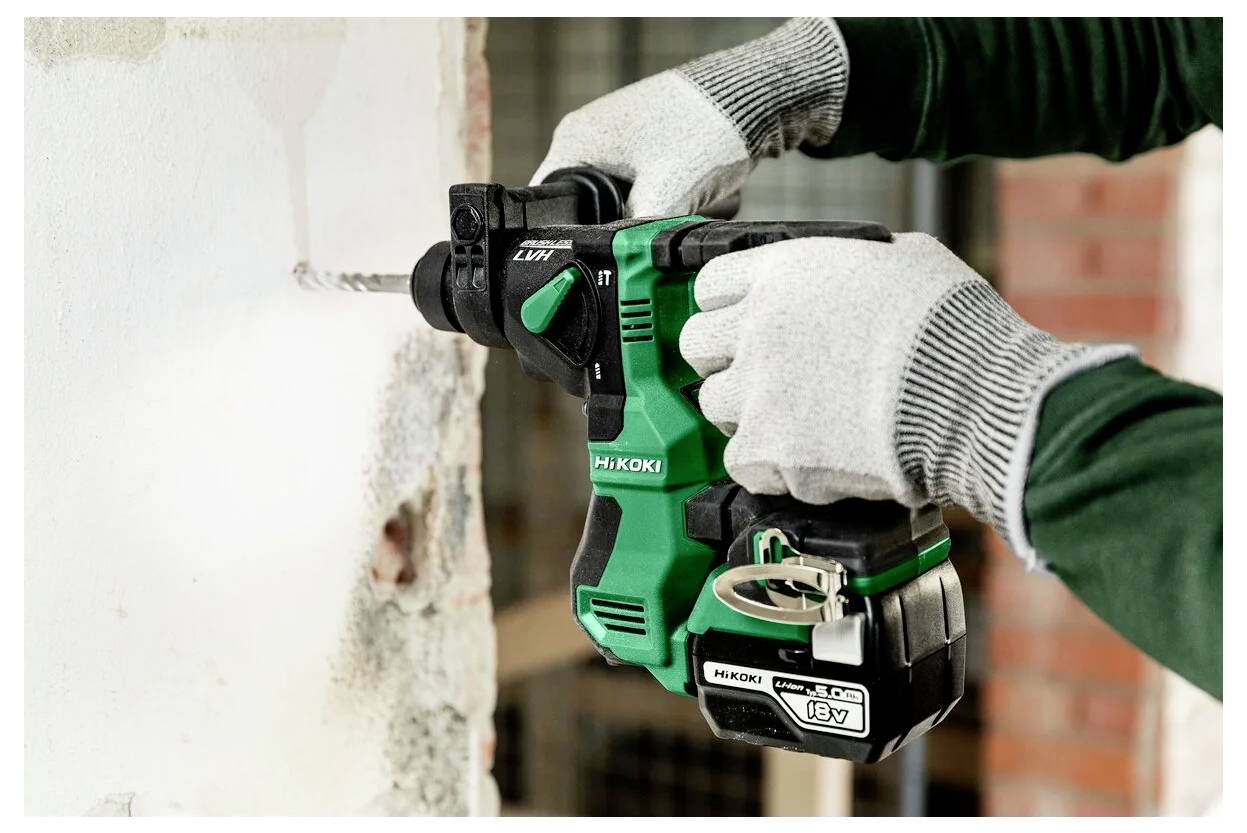 Osoba w rękawicach używająca zielonej i czarnej wiertarki akumulatorowej do wykonania otworu w białej ścianie.