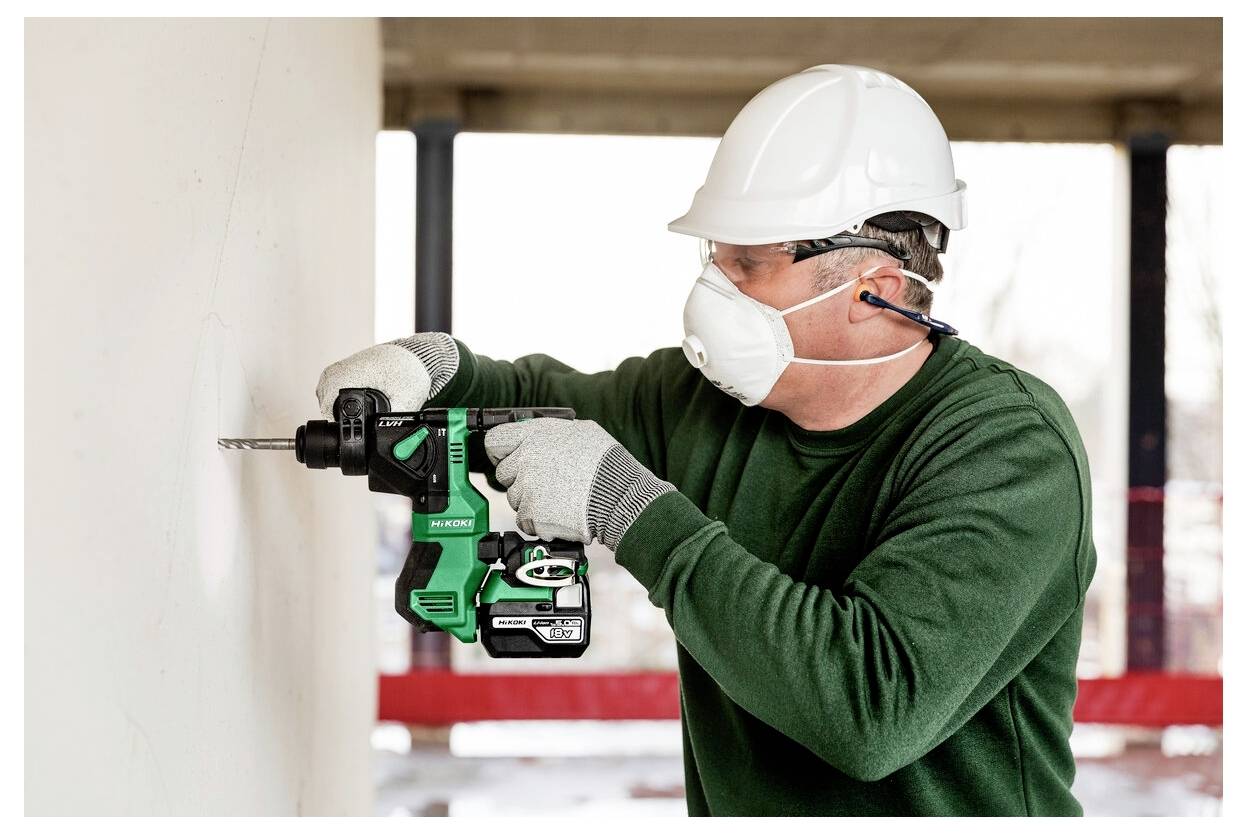 Robotnik budowlany w sprzęcie ochronnym używa zielonej wiertarki na ścianie. Pracownik nosi kask, maskę, rękawice i okulary ochronne.