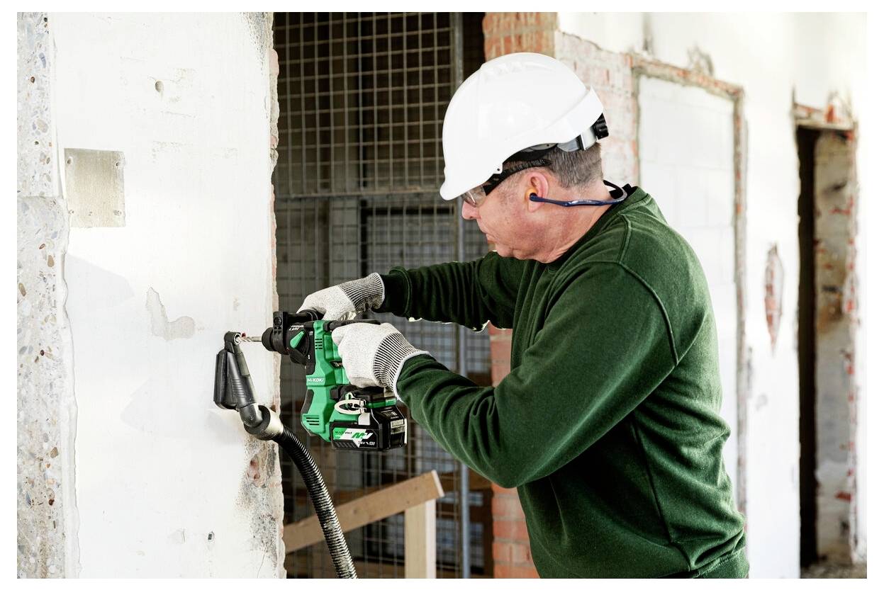 Robotnik budowlany w białym kasku ochronnym i zielonej koszuli używa wiertarki elektrycznej na ścianie w budynku będącym w trakcie remontu.