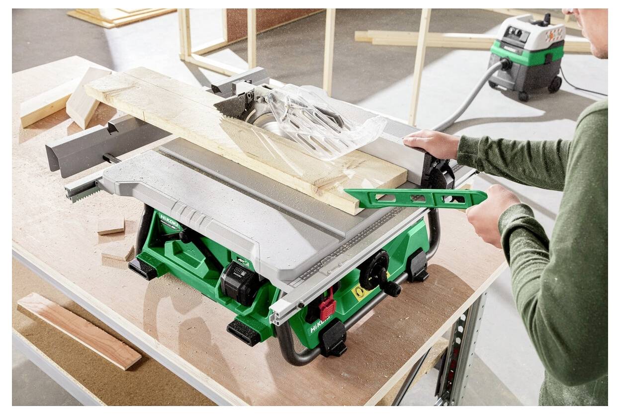 Osoba używająca zielonej i srebrnej piły stołowej do cięcia drewnianych desek. Kawałki drewna są rozrzucone dookoła, a odkurzacz znajduje się w tle.