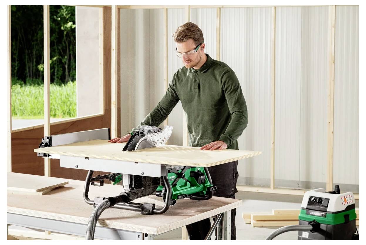 Osoba w okularach ochronnych obsługuje piłę stołową wewnątrz częściowo zbudowanego budynku, tnie drewnianą deskę.