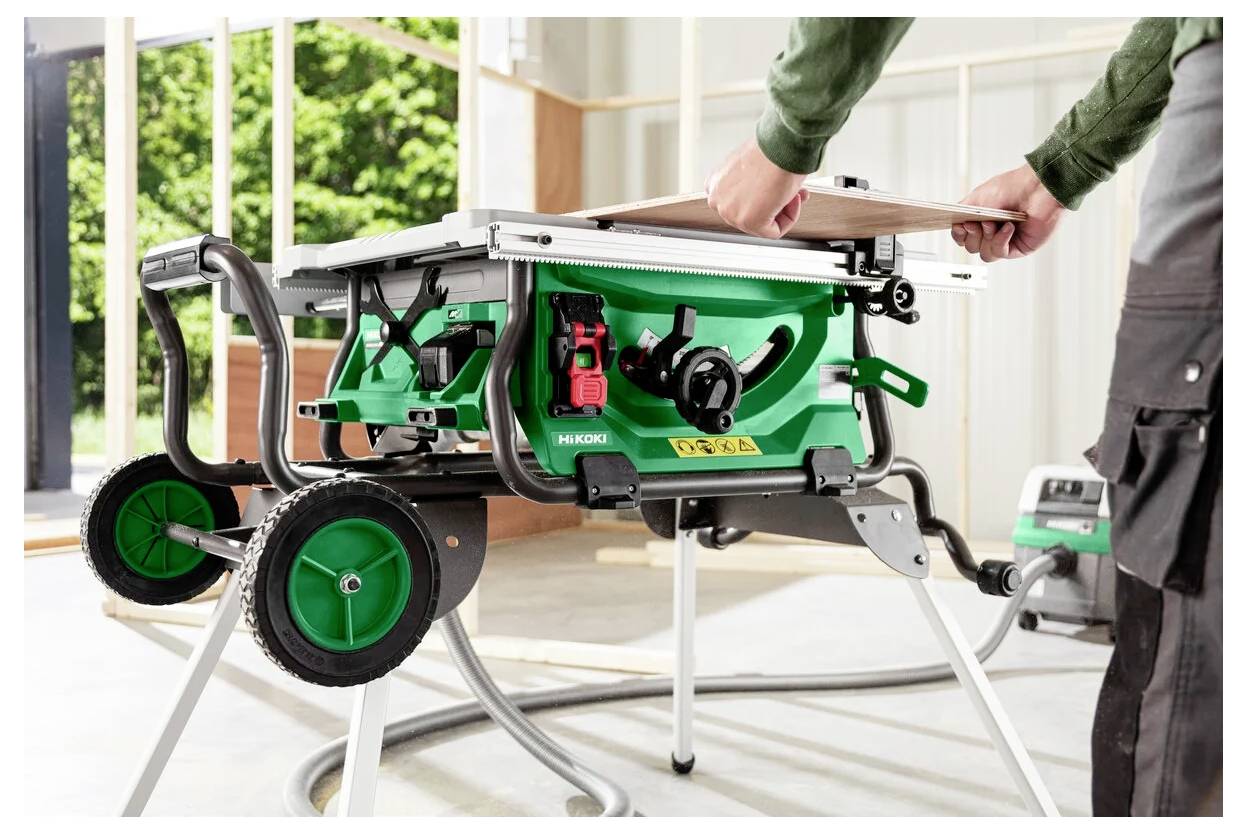 Osoba reguluje piłę tarczową w dobrze oświetlonym warsztacie. Piła jest zielona, posiada włącznik bezpieczeństwa i stoi na stojaku z kółkami.