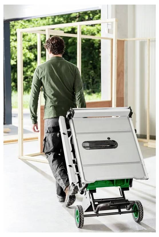 Osoba w stroju roboczym przewozi kompaktową, składaną piłę stołową na wózku w dobrze oświetlonym warsztacie, w tle widać drewniane ramy.