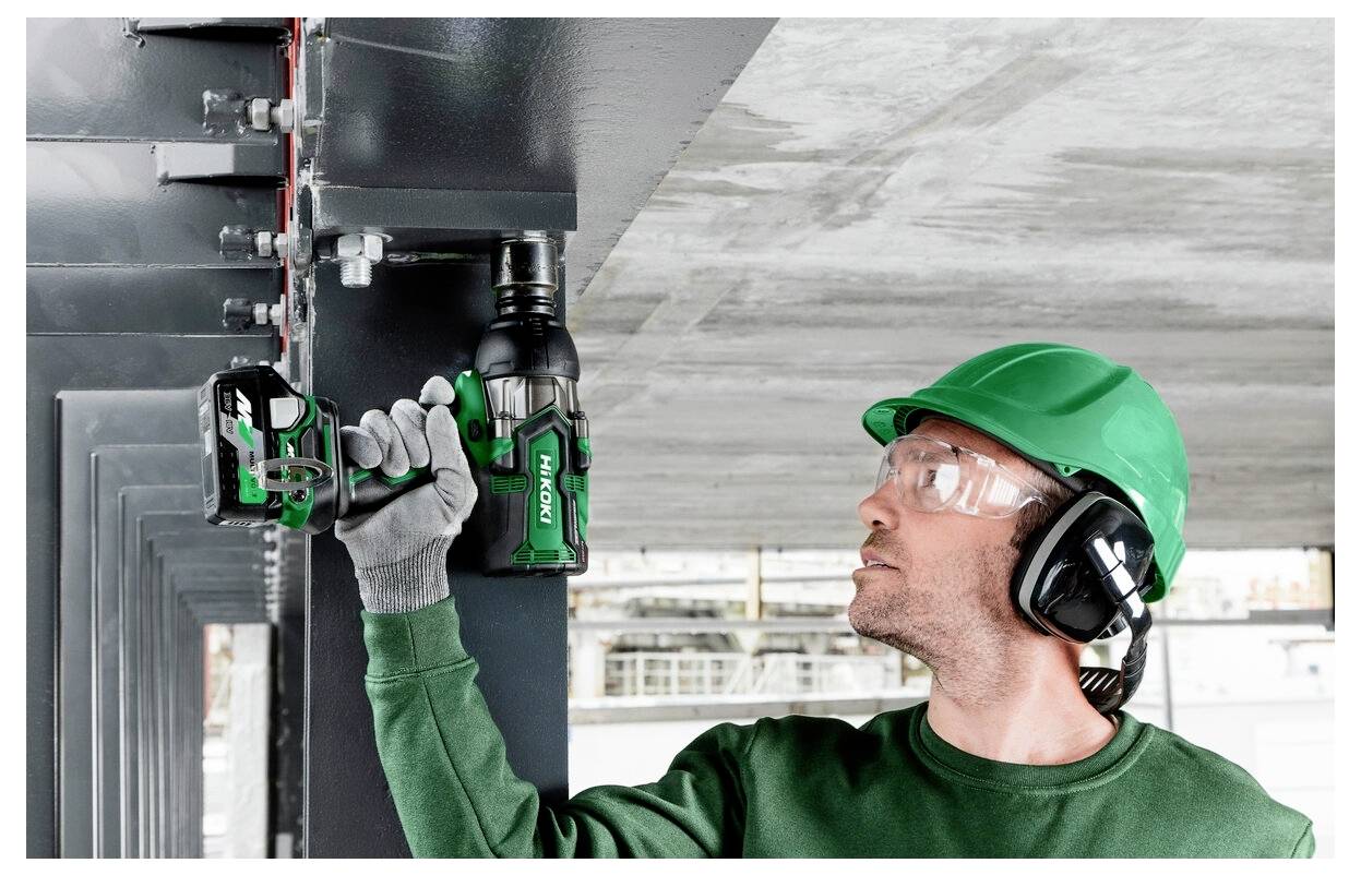 Pracownik w odzieży ochronnej używa elektronarzędzia do dokręcania śrub na stalowej belce, co wskazuje na prace budowlane lub konserwacyjne.