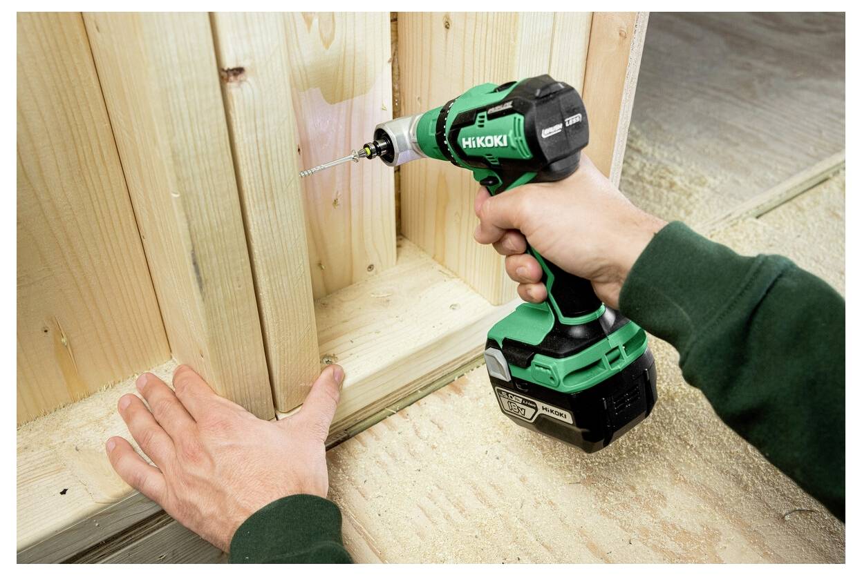 Osoba używa zielonej i czarnej wiertarki elektrycznej do wywiercenia otworu w drewnianej desce, jednocześnie przytrzymując deskę jedną ręką.