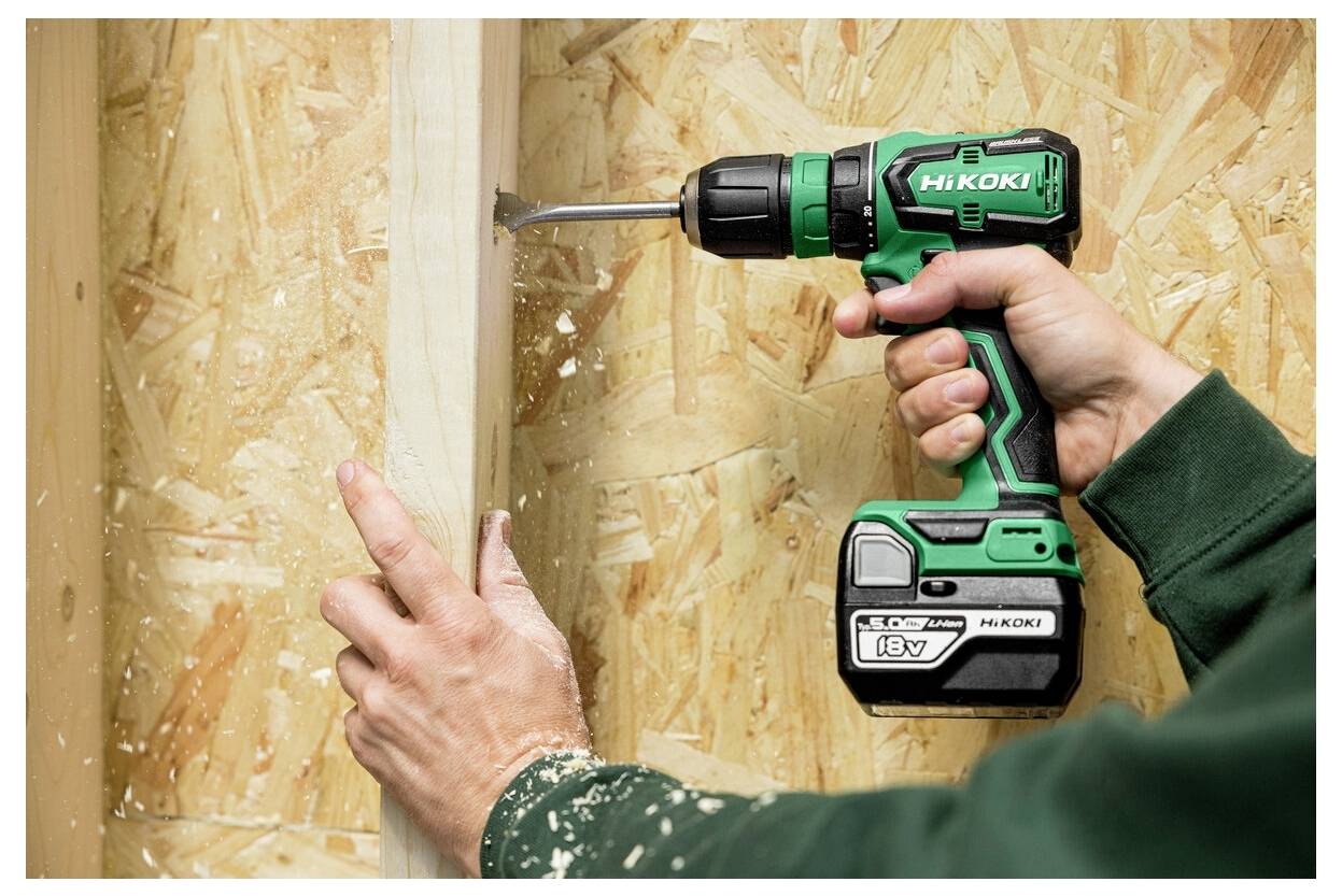Osoba używa zielonej akumulatorowej wiertarki z zamocowanym wiertłem, aby wywiercić otwór w pionowej drewnianej belce, otoczonej sklejką.