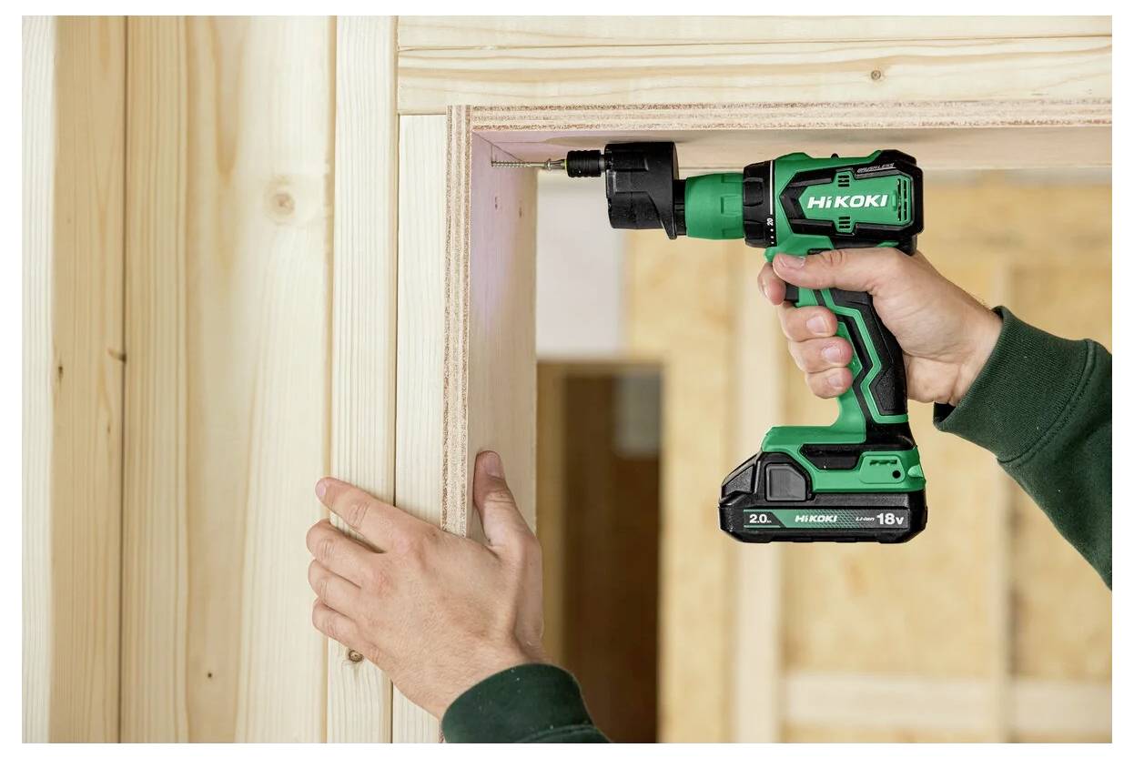 Osoba używa zielonej bezprzewodowej wiertarki do zainstalowania drewnianej futryny drzwiowej w domu w trakcie budowy. Wiertarka jest oznaczona logo 'HIKOKI'.