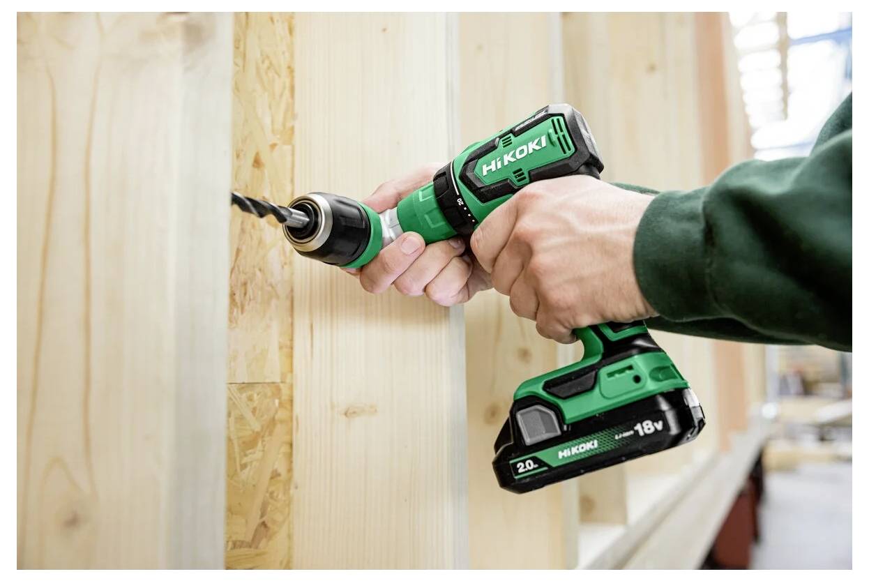 Osoba ubrana w zielony sweter używa akumulatorowej wiertarki HiKOKI do wykonania otworu w drewnianych deskach, prezentując moc i wydajność narzędzia.