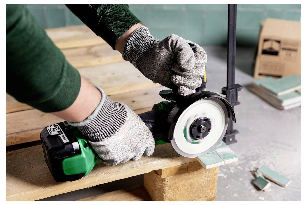 Osoba w rękawicach używa zielonego elektronarzędzia do cięcia płytki na drewnianym palecie, w pobliżu znajduje się stos płytek i zacisk.