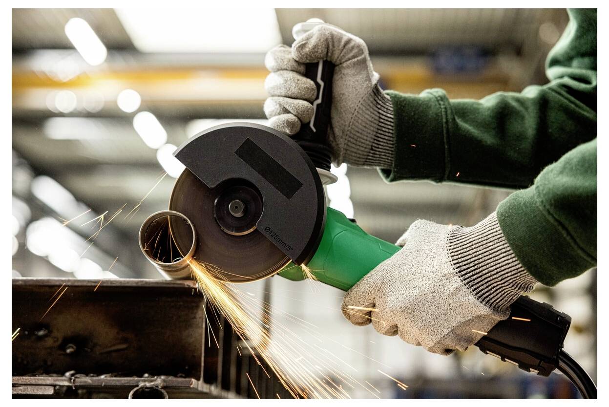 Osoba używająca szlifierki kątowej, tworząca iskry podczas cięcia metalowej rury w warsztacie. Widoczne są rękawice i sprzęt ochronny.