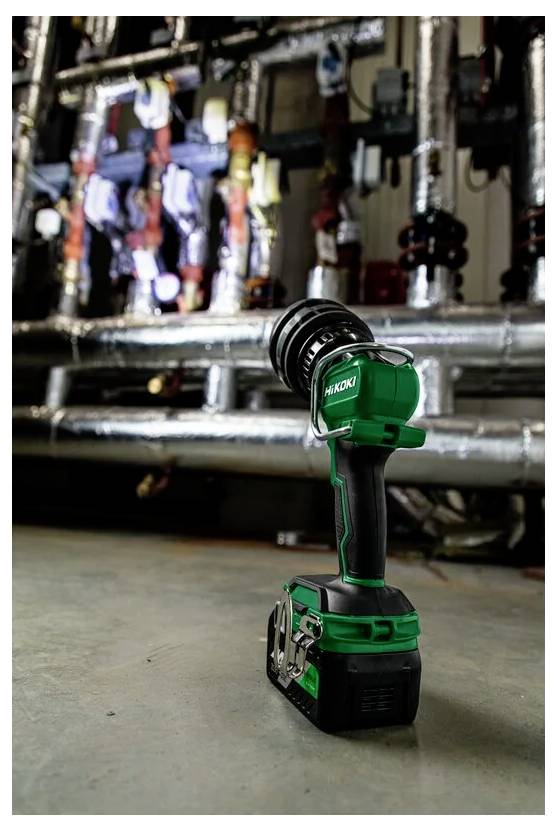 Zielona, bezprzewodowa wiertarka ręczna z czarnym uchwytem spoczywa na betonowej podłodze, a w rozmytym tle widoczne są przemysłowe rury i zawory.