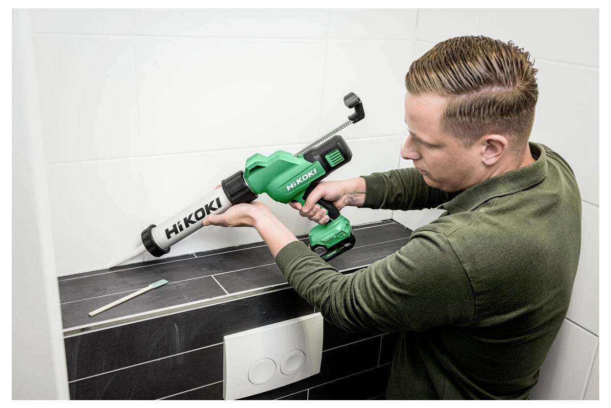 Osoba nakłada uszczelniacz na płytki łazienkowe za pomocą zielonego pistoletu do uszczelniania marki Hikoki, obok którego widoczna jest szczoteczka do zębów.