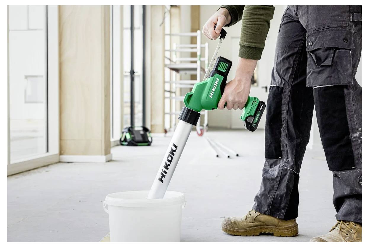 Osoba używa zielonego i białego bezprzewodowego narzędzia elektrycznego do mieszania zawartości w wiadrze wewnątrz częściowo wykończonego pomieszczenia w trakcie budowy.