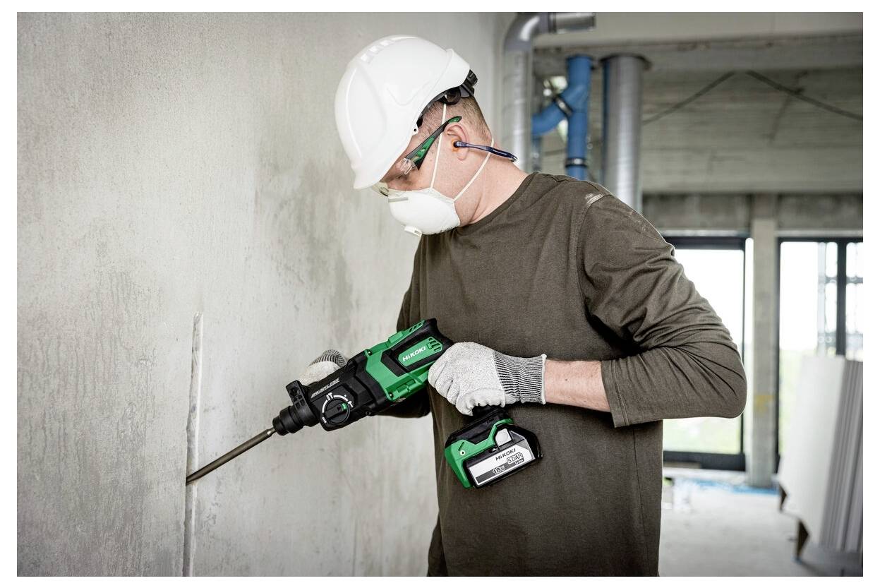 Pracownik w sprzęcie ochronnym używa zielonego elektronarzędzia do kucia betonowej ściany w częściowo wybudowanym budynku.