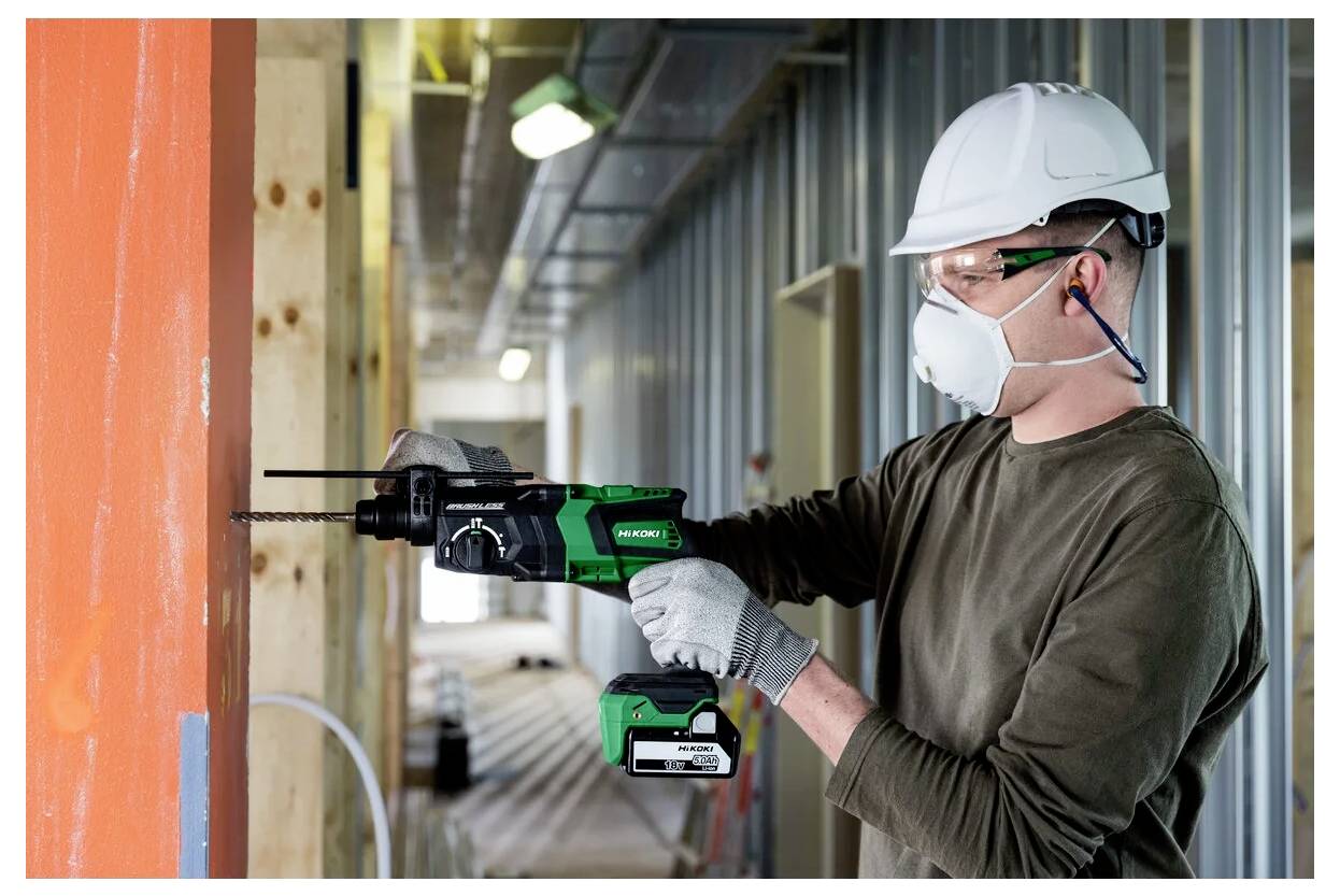 Robotnik budowlany w kasku ochronnym i stosownym sprzęcie bezpieczeństwa używa wiertarki elektrycznej na drewnianej belce w budynku będącym w trakcie budowy.