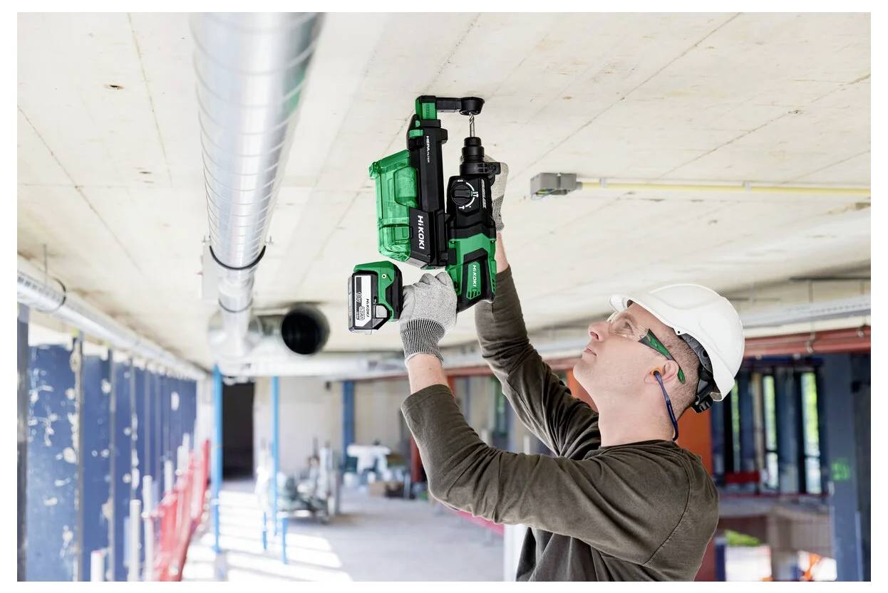 Robotnik budowlany w kamizelce ochronnej używa zielonego i czarnego elektronarzędzia, pracując przy suficie dużej, dobrze oświetlonej przestrzeni przemysłowej.