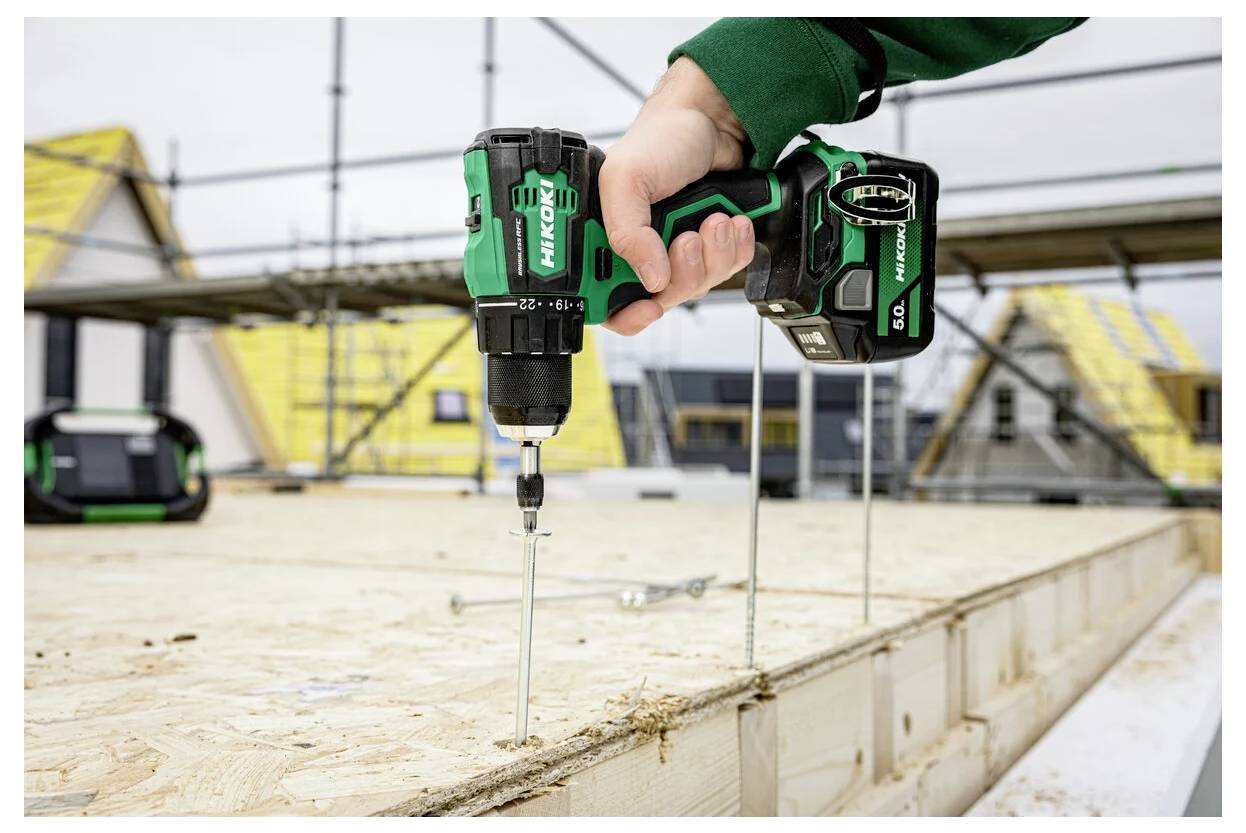 Ręka używająca zielonej bezprzewodowej wiertarki mocuje wkręt w drewnianej desce na placu budowy, z rusztowaniami i budynkami w tle.