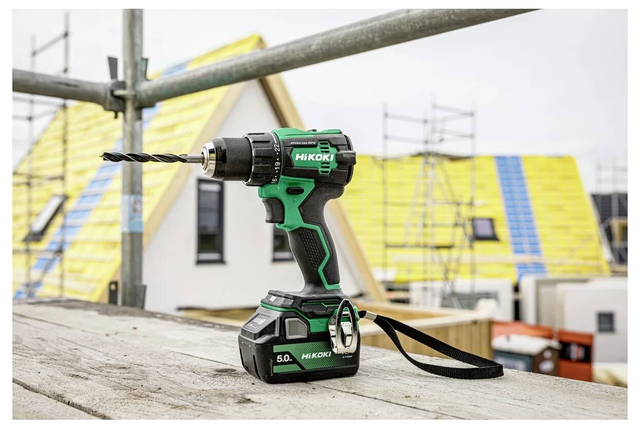 Akumulatorowa wiertarka w czarno-zielonym kolorze spoczywa na drewnianej desce na terenie budowy, w tle widać częściowo wybudowane domy.