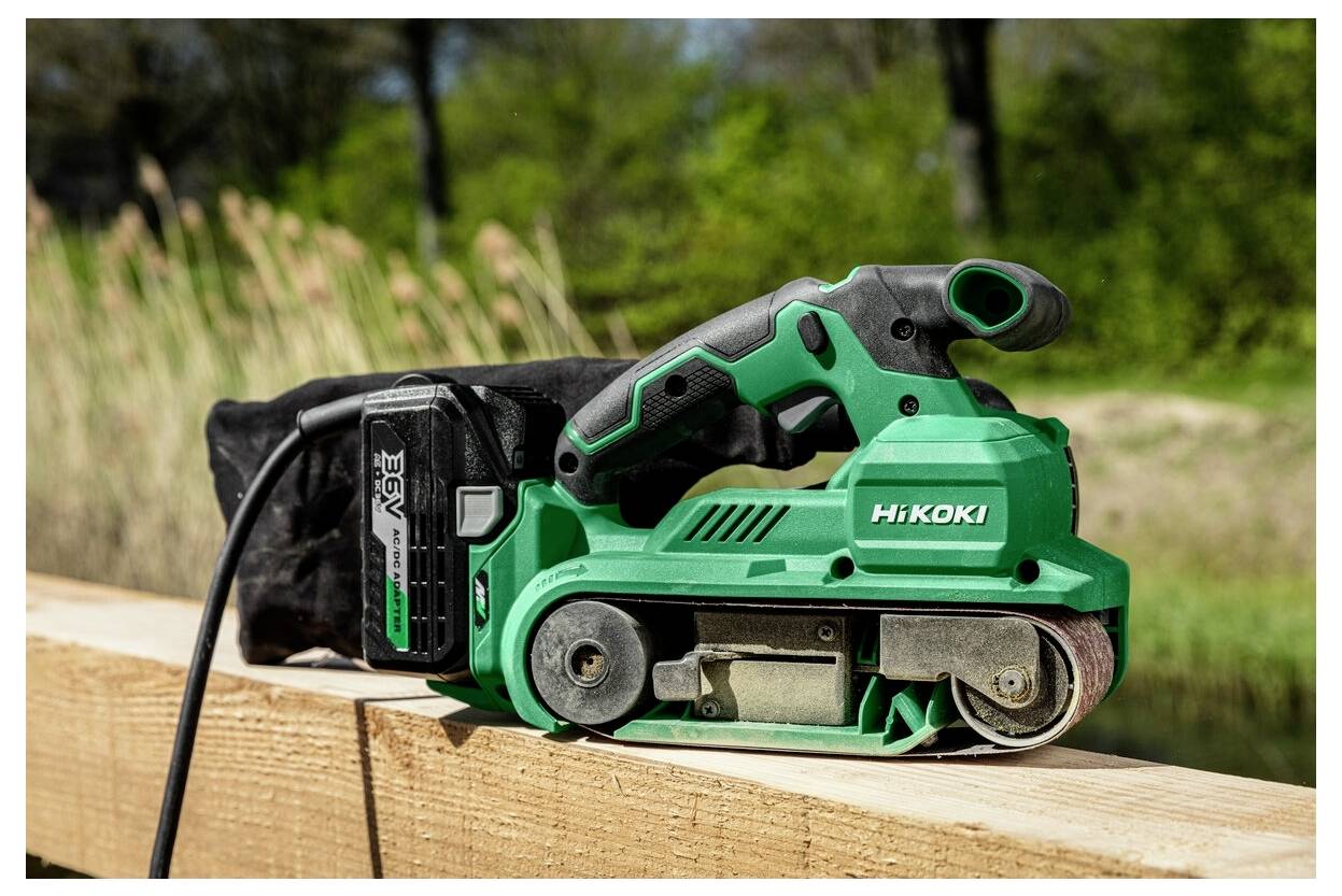 Zielona szlifierka taśmowa z czarną torbą na pył spoczywa na drewnianej desce na zewnątrz, otoczona zielenią.