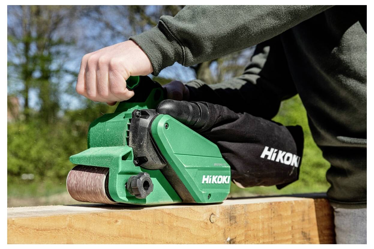 Osoba używa zielonej szlifierki taśmowej HiKOKI na drewnianej powierzchni na świeżym powietrzu, prezentując ergonomię i design narzędzia.