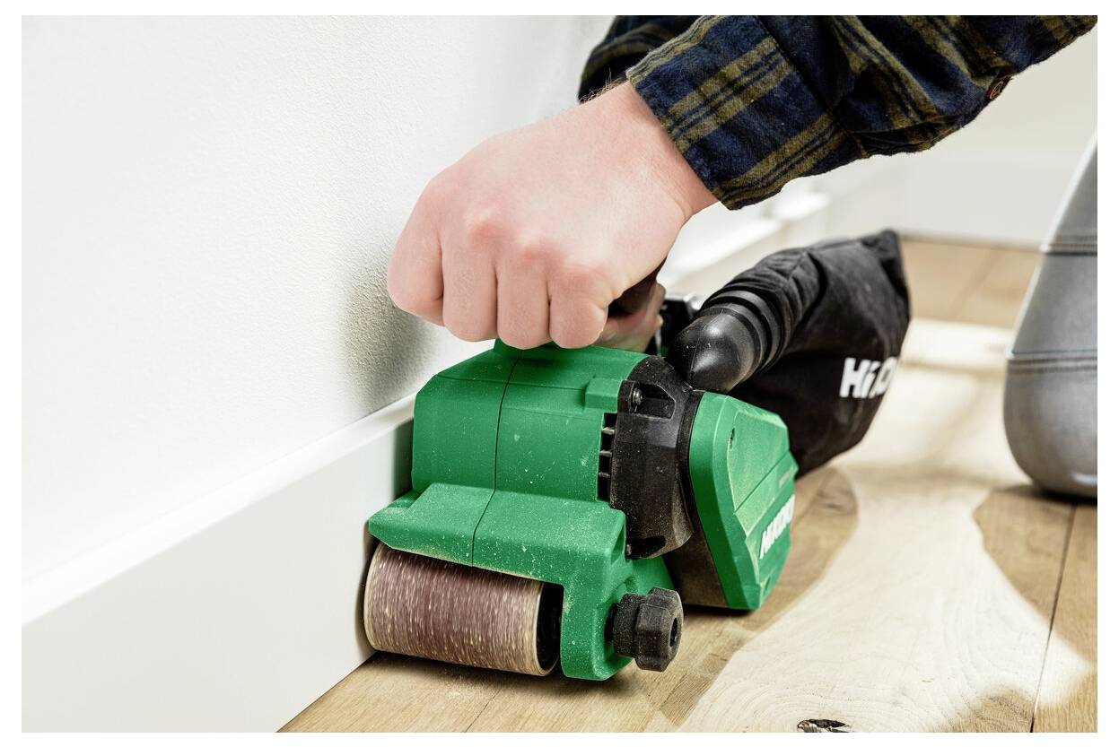 Osoba używa zielonej ręcznej szlifierki do wygładzania drewnianej podłogi w pobliżu ściany, ukazując czynność związaną z renowacją domu.