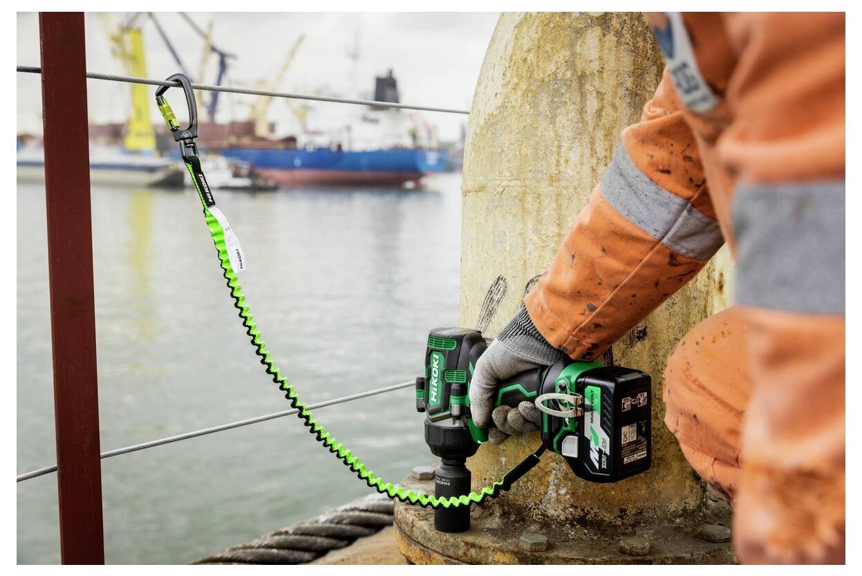 Pracownik w pomarańczowym stroju ochronnym używa wiertarki elektrycznej na metalowej konstrukcji w pobliżu nabrzeża, a w tle widoczne są statki towarowe i dźwigi.