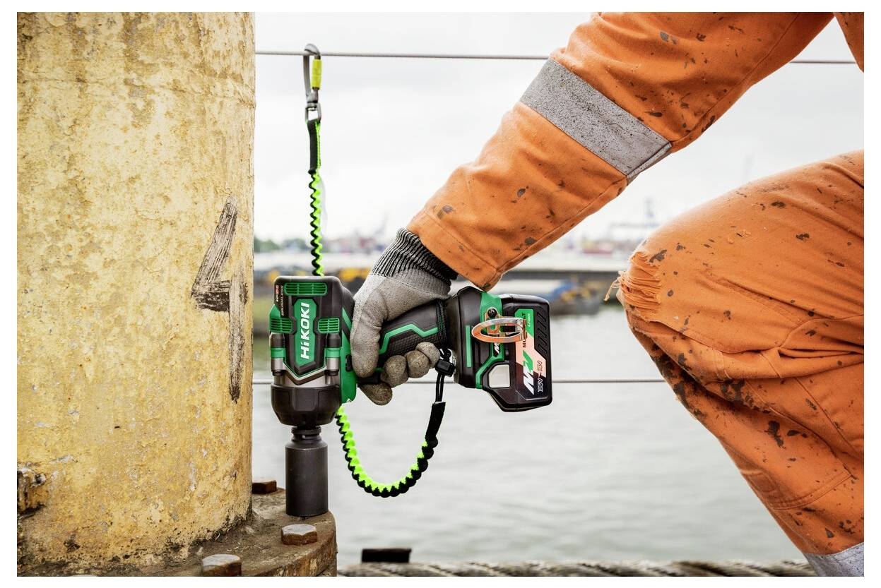 Osoba ubrana w pomarańczowy kombinezon ochronny i rękawice używa zielonego elektronarzędzia HiKOKI do pracy na dużej metalowej strukturze nad wodą.