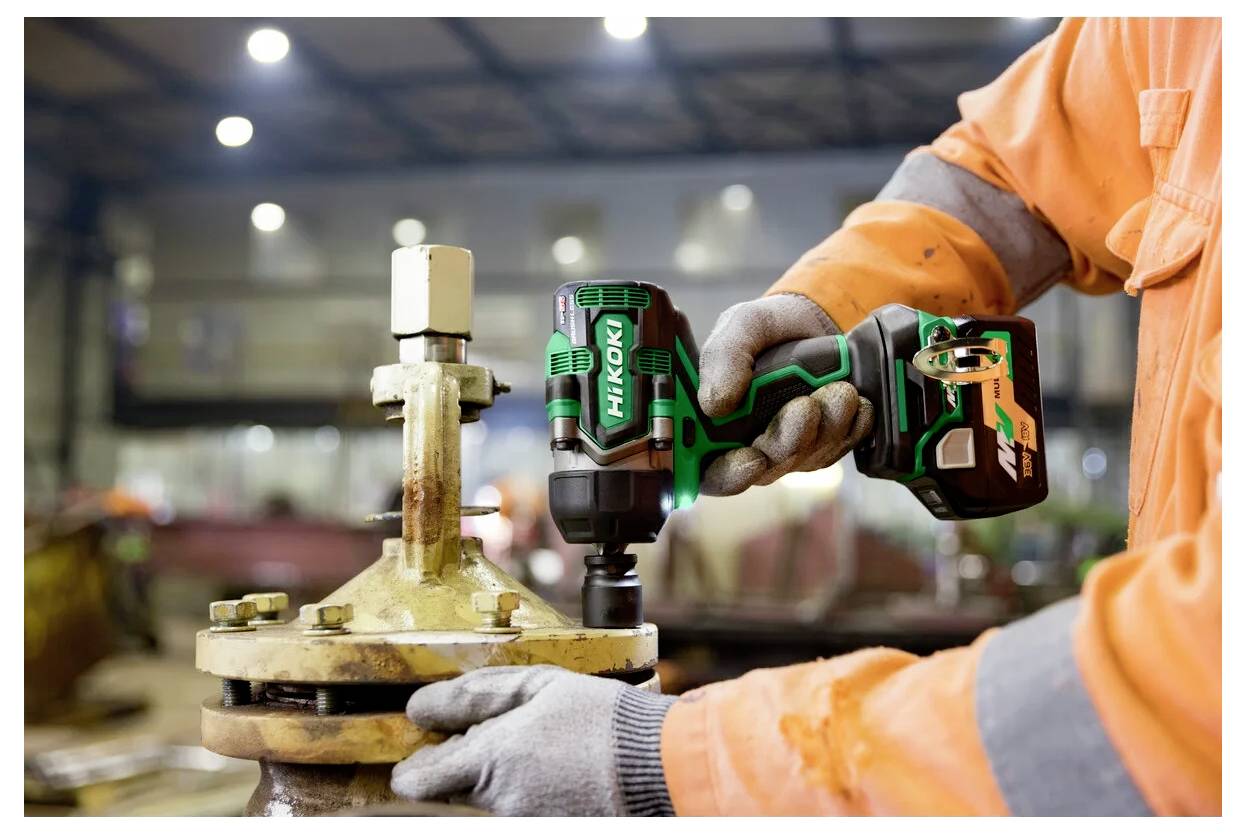 Osoba ubrana w pomarańczową roboczą kurtkę używa elektronarzędzia HiKOKI do dokręcenia śruby na sprzęcie przemysłowym wewnątrz warsztatu.
