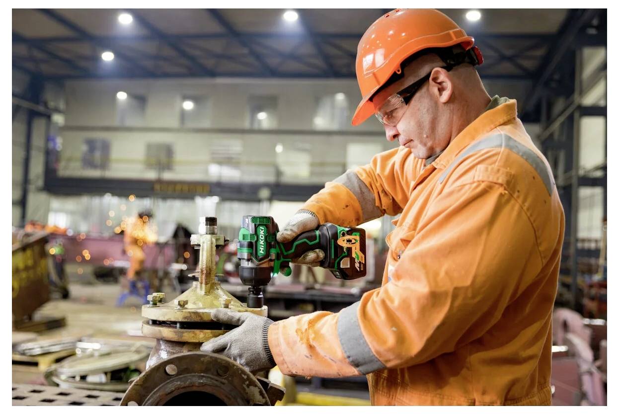 Pracownik w pomarańczowym kombinezonie ochronnym i kasku używa wiertarki elektrycznej na przemysłowej maszynie w fabrycznym otoczeniu, działając z koncentracją i precyzją.