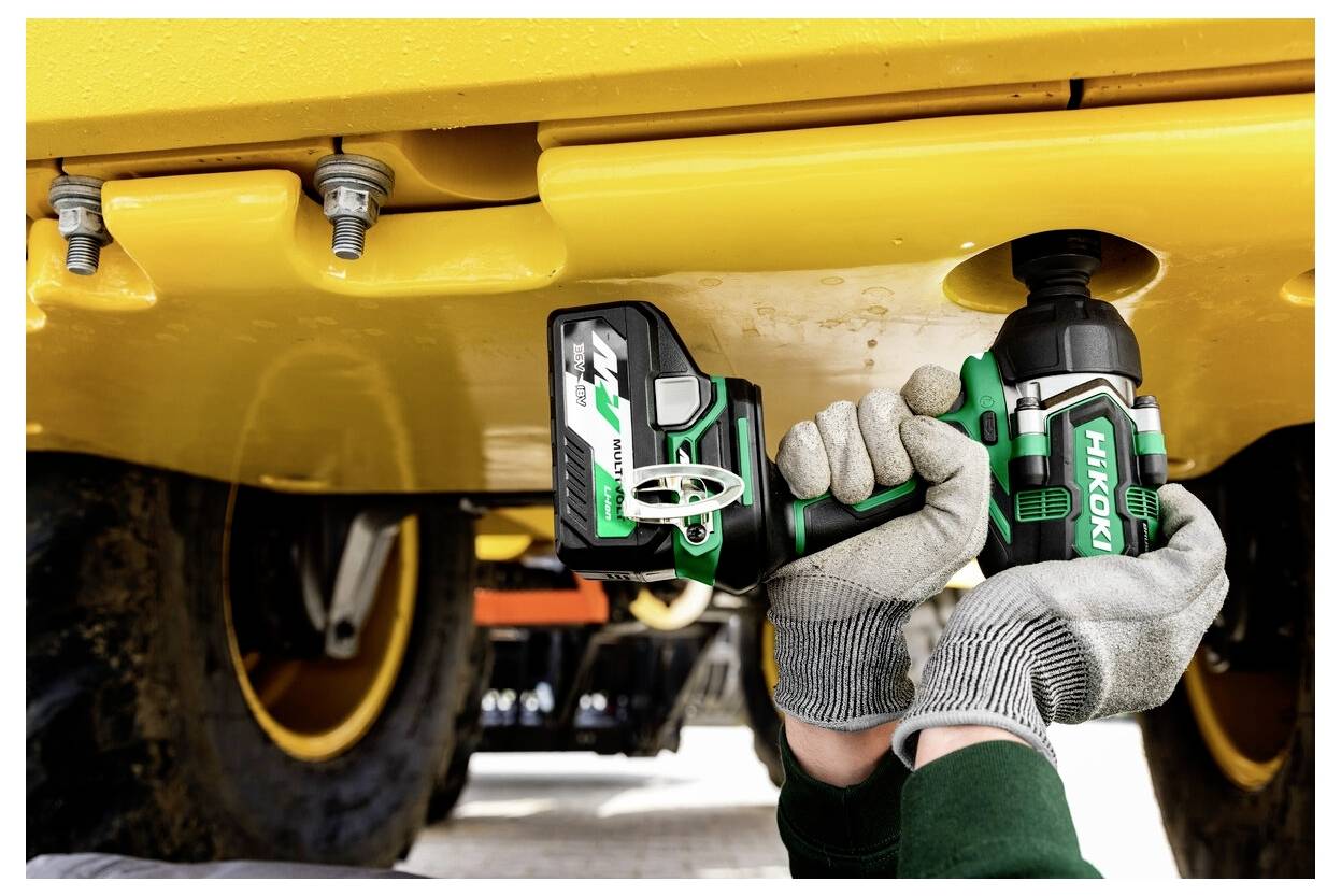 Osoba używa zielonego i czarnego narzędzia elektrycznego do dokręcania śruby pod ciężkim sprzętem przemysłowym, mając na sobie rękawice ochronne.