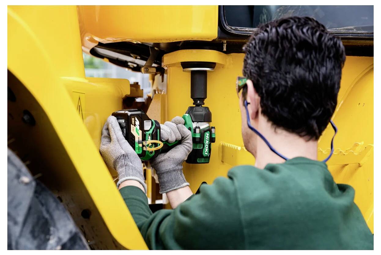 Osoba nosząca rękawice i ochronniki słuchu używa zielonej bezprzewodowej wiertarki do pracy przy dużej żółtej maszynie, prawdopodobnie podczas konserwacji.