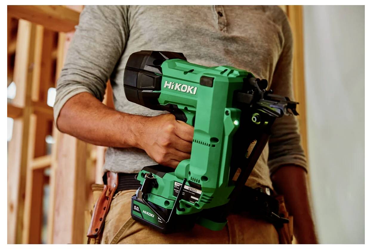 Osoba w odzieży roboczej trzyma zielony pistolet do gwoździ HiKOKI na placu budowy, a w tle widoczne są drewniane ramy.