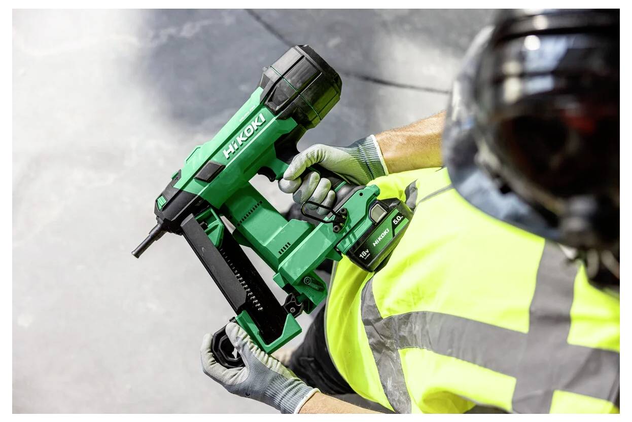 Pracownik w kamizelce ostrzegawczej trzymający zielone narzędzie elektryczne Hitachi, prawdopodobnie gwoździarkę, w otoczeniu przemysłowym.