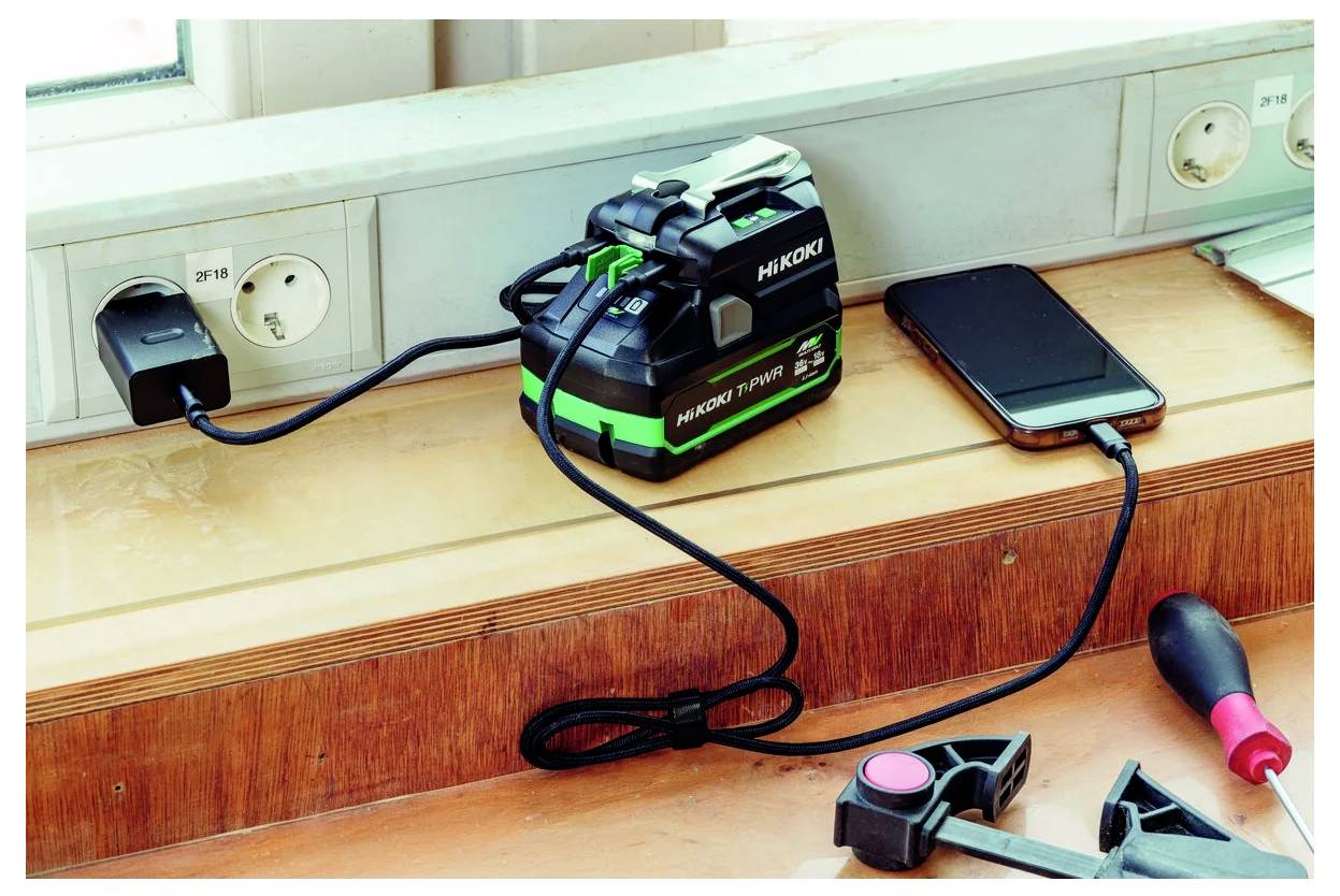Bateria podłączona do listwy zasilającej oraz smartfon na drewnianej półce, obok leży śrubokręt i narzędzie.
