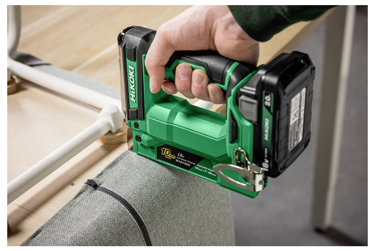 Osoba używa zielonego, bezprzewodowego zszywacza Hikoki do przymocowania tkaniny do drewnianej ramy na stole.