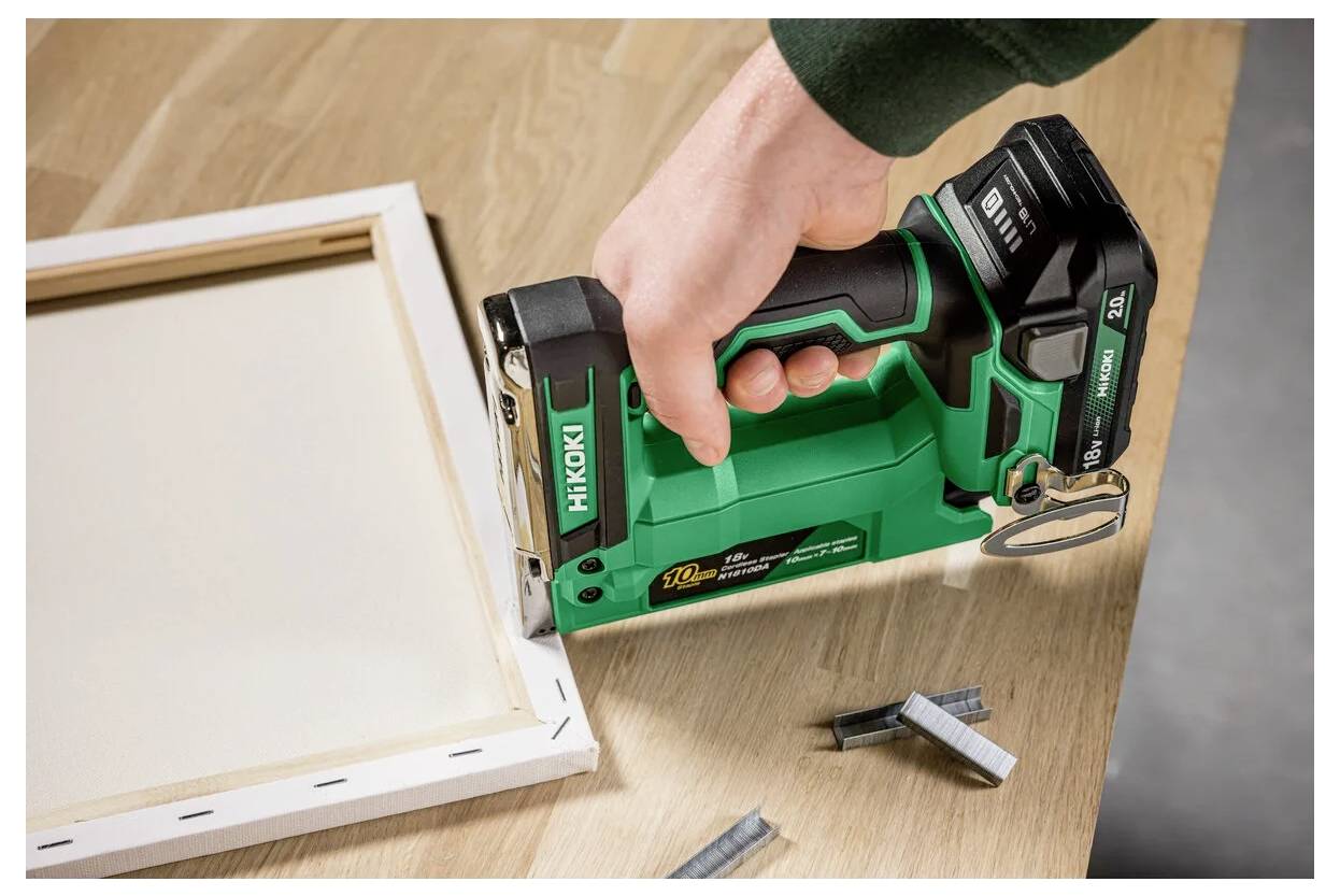 Osoba używa zielonego i czarnego elektrycznego zszywacza do przymocowania ramy płótna. Kilka zszywek jest rozrzuconych na drewnianej powierzchni.