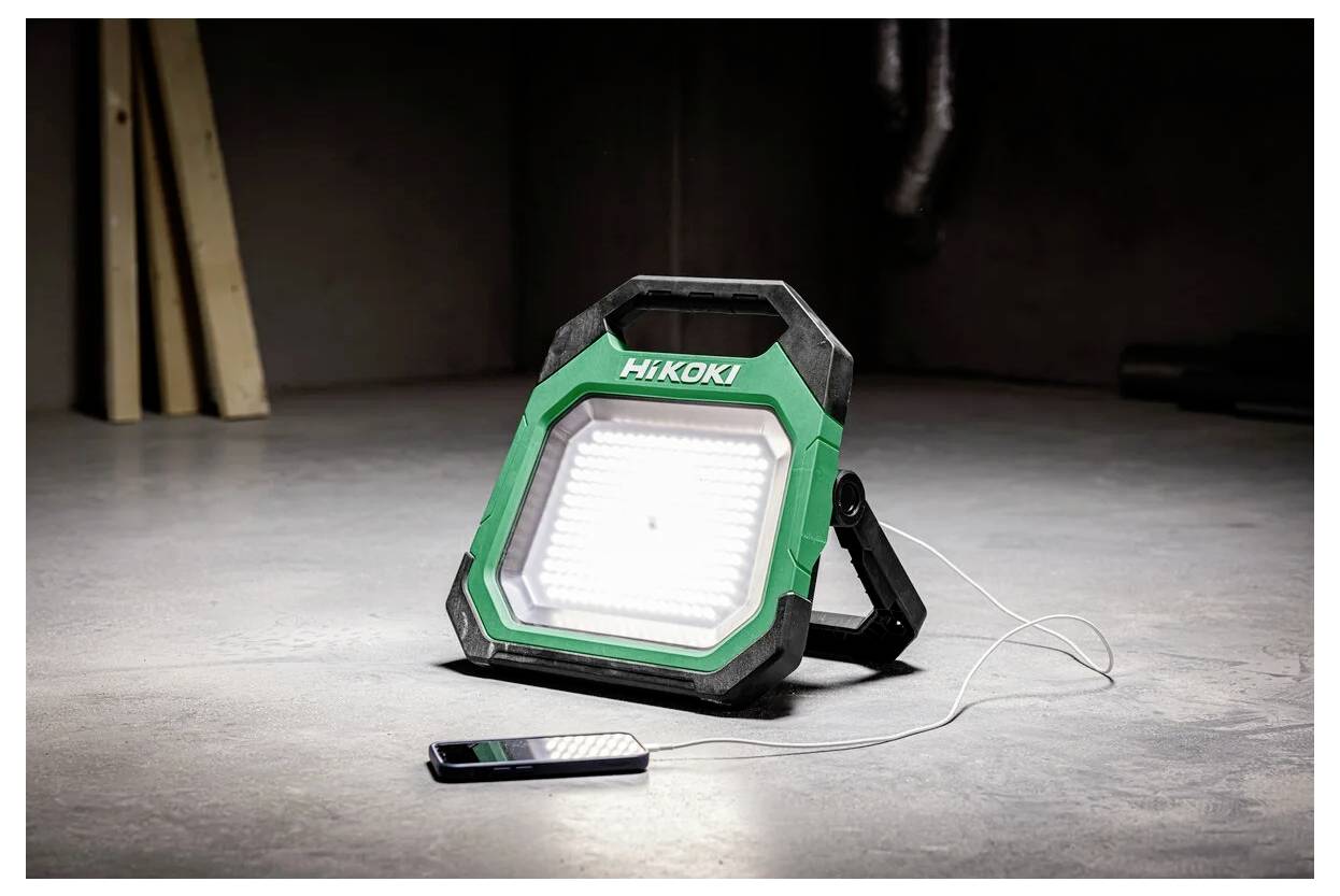 Zielono-czarna, przenośna lampa robocza LED z etykietą 'HiKOKI' jest ustawiona na podłodze, oświetlając okolicę. Obok niej ładuje się smartfon.
