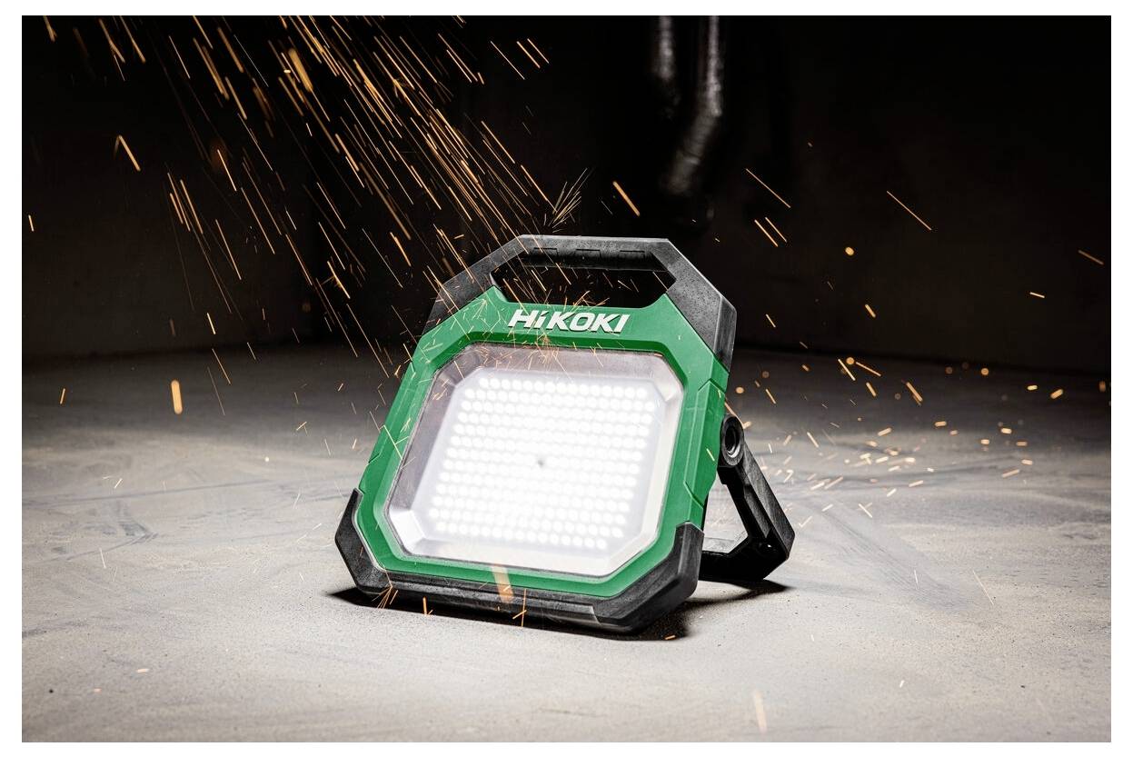 Przenośna lampa robocza oznaczona logo 'HiKOKI' w warsztacie, emitująca jasne światło z widocznymi iskrami latającymi wokół, sugerująca aktywne użycie.