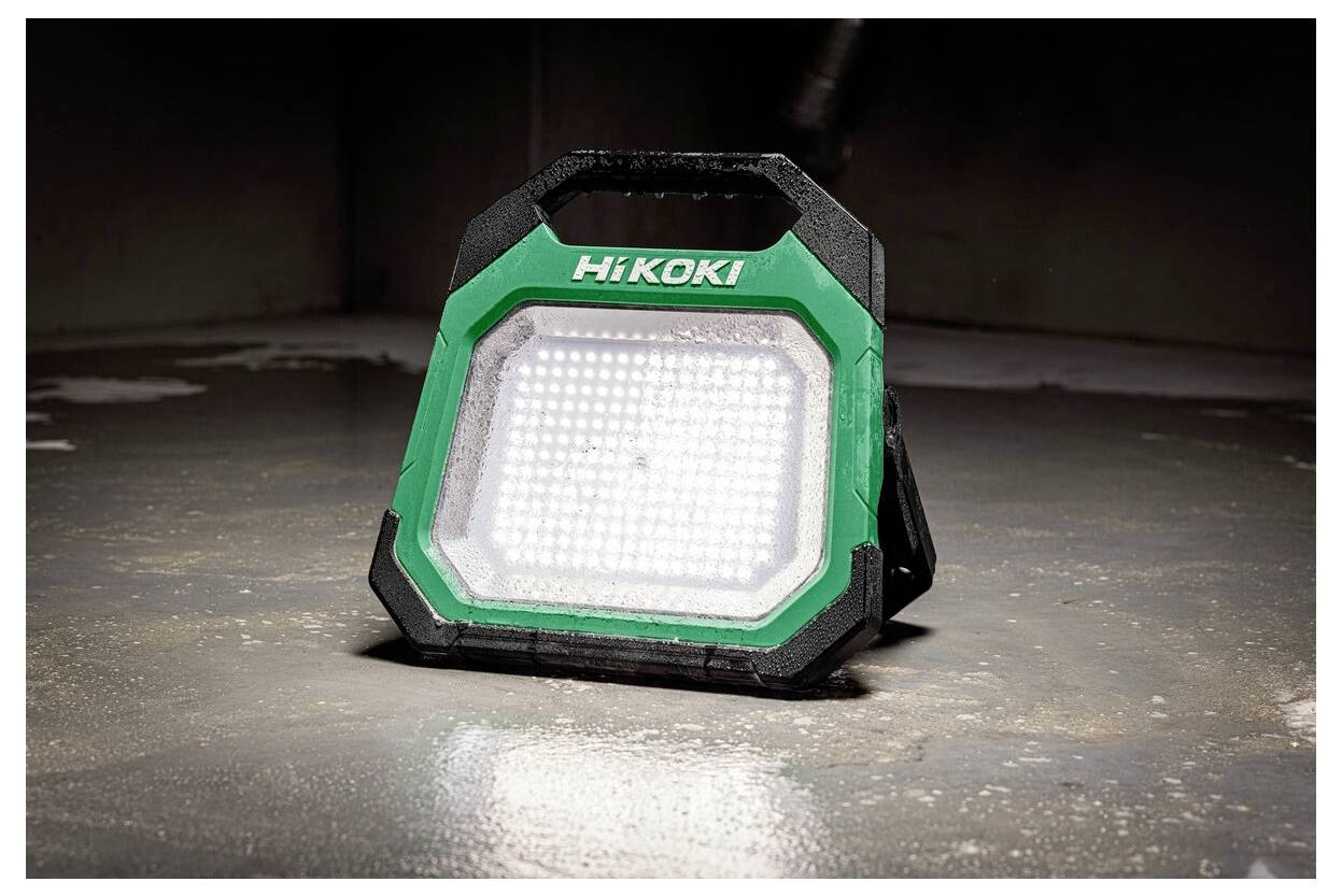 Zielona i czarna przenośna lampa robocza LED marki 'HiKOKI' stoi na betonowej podłodze, oświetlając słabo oświetlony obszar.