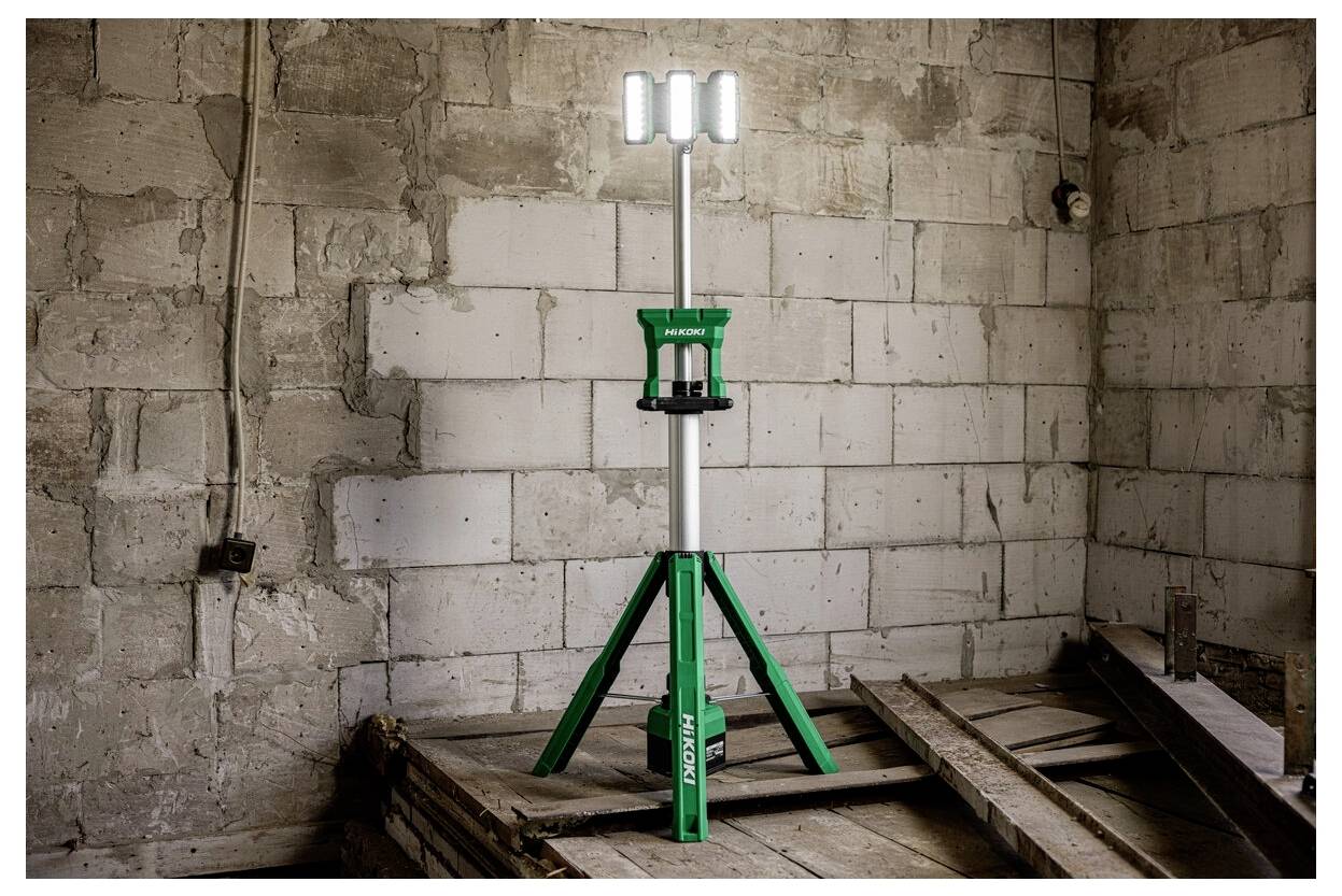 Zielona lampa budowlana zamontowana na trójnogu stoi w częściowo wybudowanym pomieszczeniu o nagich ceglanych ścianach, zapewniając przenośne oświetlenie terenu budowy.