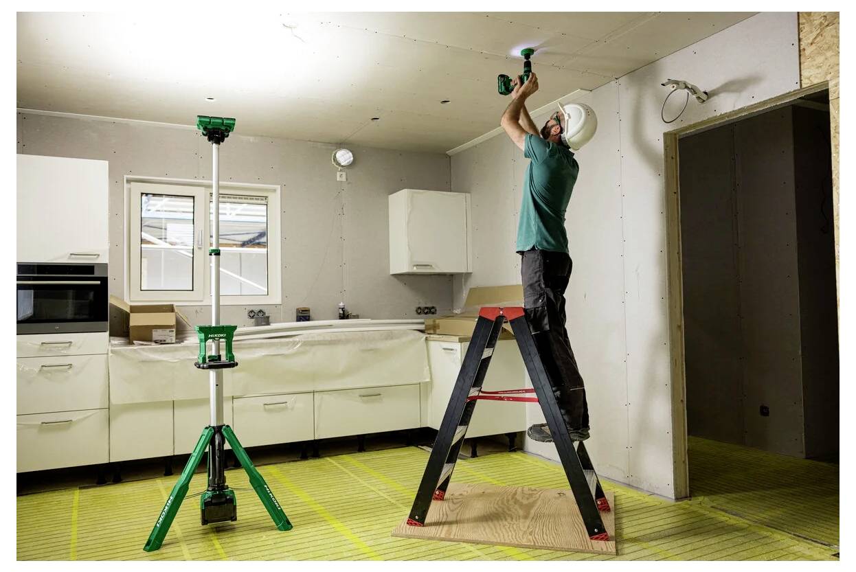Osoba stoi na drabinie, używając elektronarzędzia do przymocowania czegoś do sufitu w częściowo urządzonej kuchni z białymi szafkami.