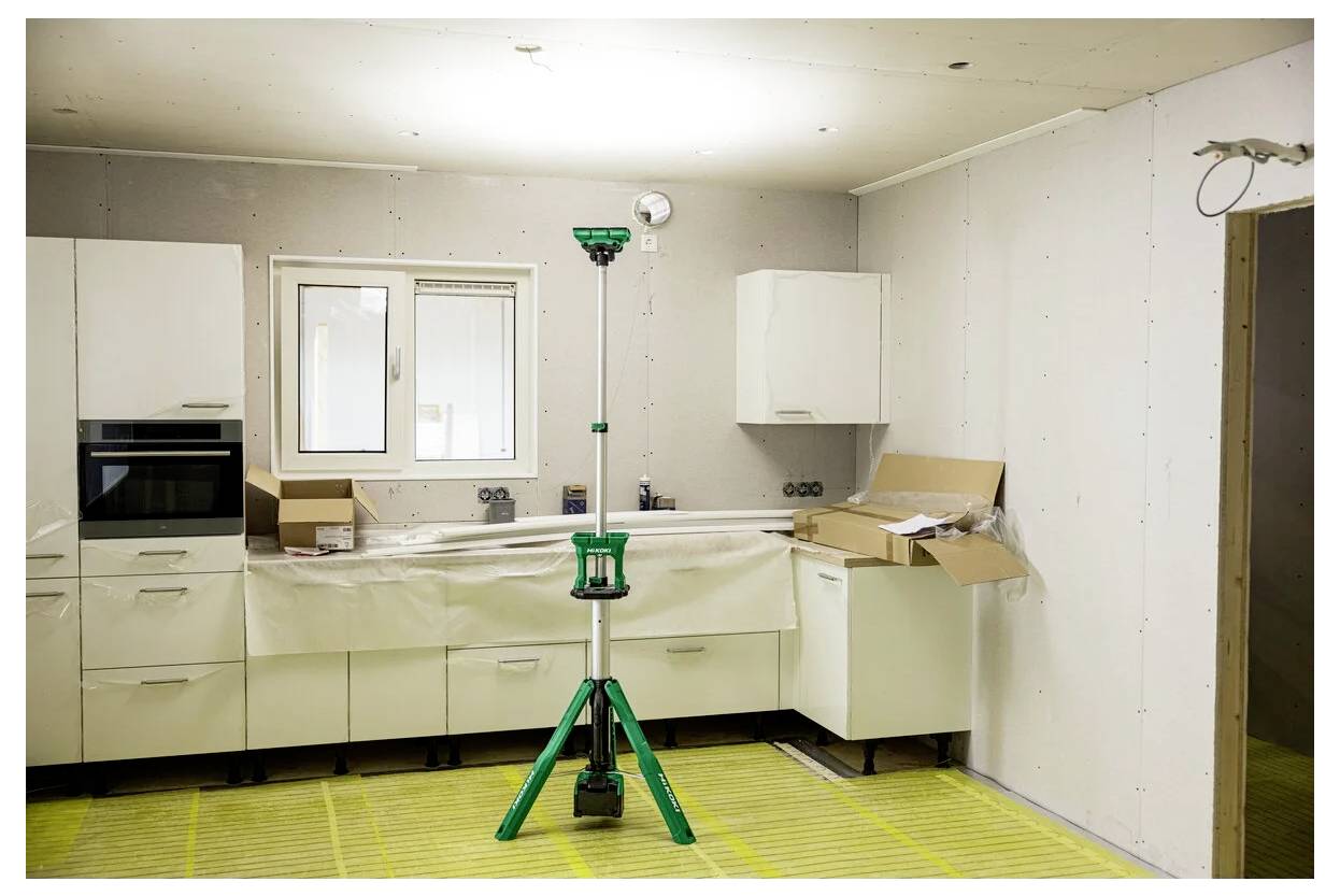 Kuchnia w trakcie remontu z białymi szafkami, oknem i narzędziami budowlanymi. Na podłodze znajdują się kartony i sprzęt.