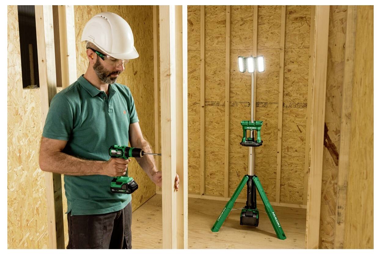 Osoba w ubraniu ochronnym używa wiertarki wewnątrz drewnianej struktury ramowej, przy świetle roboczym na statywie oświetlającym przestrzeń.