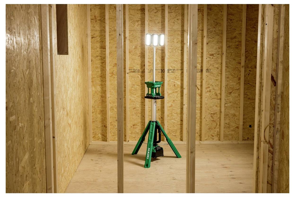 Przenośna lampa robocza stoi w centrum pomieszczenia będącego w trakcie budowy, z izolowanymi ścianami z płyt pilśniowych i widocznymi drewnianymi elementami konstrukcyjnymi.