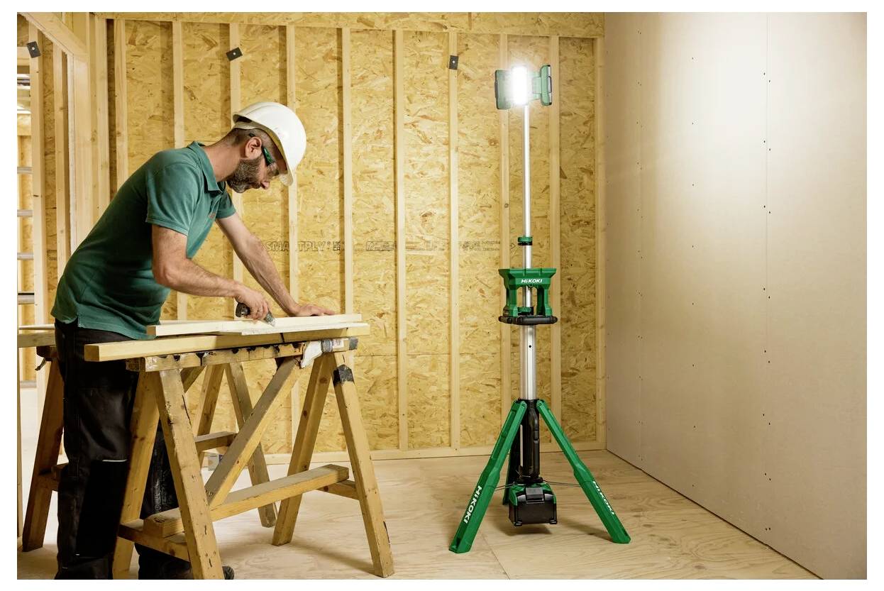Mężczyzna w kasku mierzy drewnianą deskę na kobyłce stolarskiej w częściowo wybudowanym pomieszczeniu, oświetlonym przez stojącą lampę roboczą.