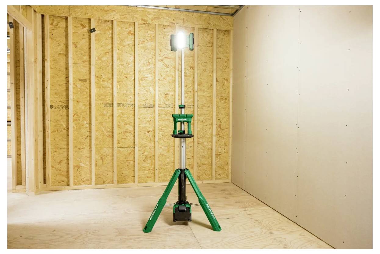 Zielony statyw z lampą roboczą jest ustawiony w częściowo wykończonym pokoju, gdzie po lewej stronie znajdują się drewniane ściany, a po prawej białe ściany.